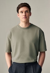 Mellemvægt olivengrøn bomuld T-shirt med korte ærmer, rund hals og subtil struktur, kombineret med mørke skræddersyede bukser.