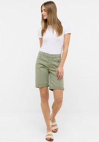 Shorts vert olive en mélange de coton, avec une coupe classique, des revers roulés et des poches avant, associés à un t-shirt blanc et des sandales.