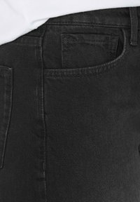 Schwarze Jeans aus Denim mit schlichtem Design, seitlicher Tasche und glatter Textur. Die Nähte sind entlang der Säume sichtbar.