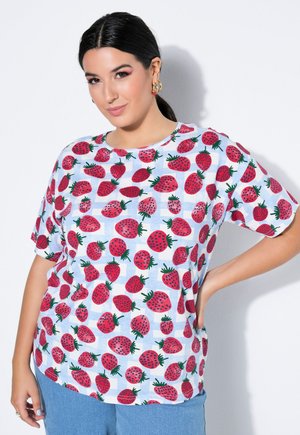 Vrouw met donker haar in een paardenstaart die gouden creolen draagt en een wit overhemd met rode aardbeienpatronen, gecombineerd met lichtblauwe spijkerbroek.