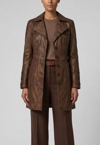 Kvinde iført en brun læderknapfrakke over en brun sweater og brune bukser, stående mod en ensfarvet grå baggrund.