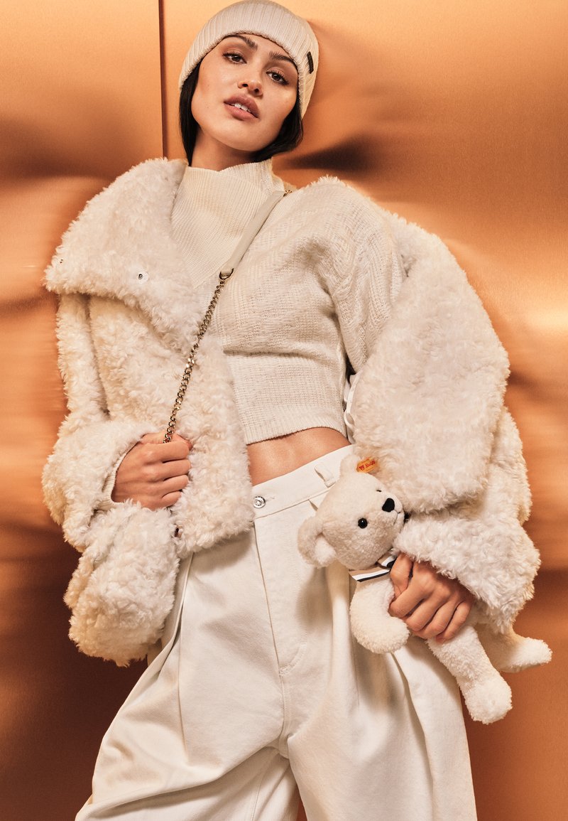 Prenda exterior de pelaje blanco con mangas extragrandes, combinada con un suéter acanalado crema y pantalones blancos sueltos. Sosteniendo un pequeño oso de peluche.