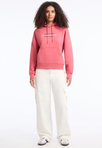 Felpa con cappuccio rosa e tasca frontale, con un logo bianco. Abbinata a pantaloni cargo a gamba larga bianco sporco e scarpe da ginnastica multicolori.