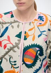 Gros plan d'une personne portant une veste crème avec une broderie florale colorée et cachemire, et un bord rose clair autour de l'encolure.