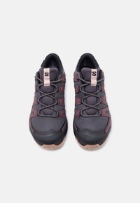 Pereche de pantofi sport gri închis și mov, din material plasă, cu șireturi elastice și tălpi maro deschis, pe un fundal alb.