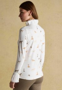 Biela košeľa s dlhým rukávom z látky s béžovými zvieracími potlačami. Má vysoký volánikový golier a manžety na gombíky s čipkovaným lemovaním.