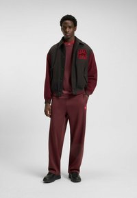 Black varsity jacket with red embroidery over a burgundy sweatshirt and matching sweatpants, featuring a relaxed fit and streamlined silhouette.