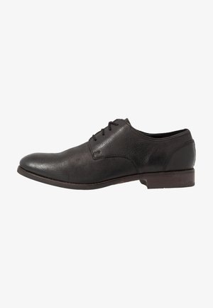 Chaussure habillée pour homme en cuir noir avec fermeture à lacets et petit talon empilé, vue de côté sur fond blanc.