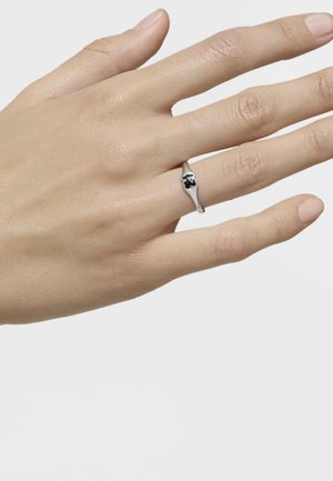 Anillo de plata con un diseño de trébol negro, montado sobre una banda delgada. Mano posicionada con los dedos ligeramente separados, contra un fondo liso.