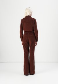 Bruine corduroy jas gecombineerd met bijpassende fluïde broek. Kenmerken zijn een western-stijl schouderpas, zakken en een gestructureerde stof. Achteraanzicht getoond.