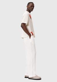 Hombre con camisa blanca de manga corta con estampados de flores rojas y pantalones anchos blancos, de pie con las manos en los bolsillos, llevando zapatos beige.