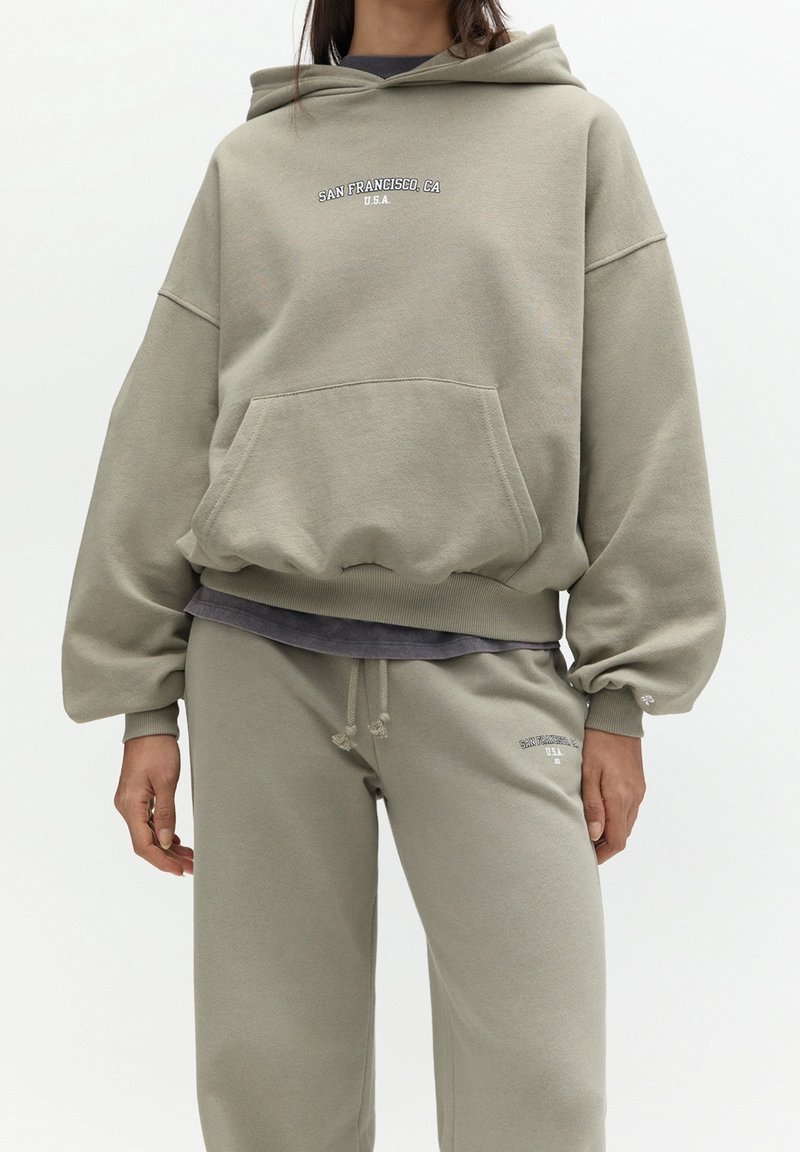 Personne portant un sweat à capuche beige ample et un pantalon de survêtement assorti avec le texte "San Francisco, CA U.S.A." sur la poitrine et la cuisse.