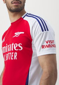 Tricou de fotbal roșu și alb cu accente albastre, având logo-ul "Emirates" în alb și textul "Visit Rwanda" pe mânecă.