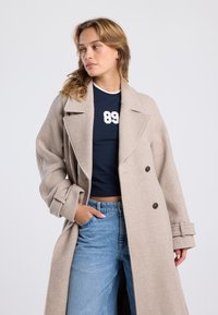 Abrigo largo de mezcla de lana color beige con solapas oversized y cierre de doble botonadura. Llevado sobre un top corto azul marino y jeans de tiro alto en un azul claro.