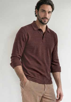 Mann mit dunklen Haaren und Bart, der ein braunes langärmeliges Poloshirt und beige Cordhosen trägt, steht mit den Händen in den Taschen.