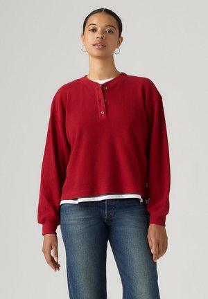 Giovane donna che indossa una camicia rossa a maniche lunghe con bottoni sopra una maglietta bianca e jeans blu, in piedi davanti a uno sfondo chiaro e semplice.