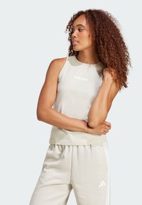 Top sin mangas de color beige claro con cuello redondo y logo blanco de "adidas"; combinado con pantalones de chándal de beige claro a juego, que presentan tres rayas blancas.