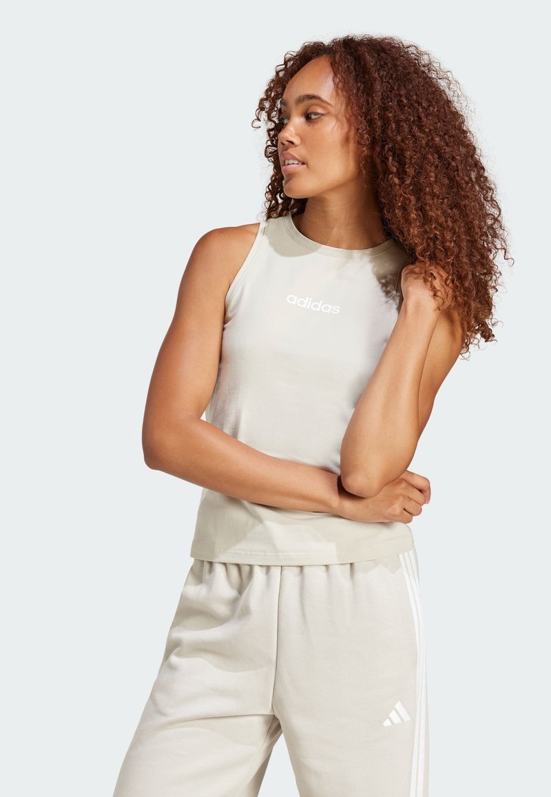 Top sin mangas de color beige claro con cuello redondo y logo blanco de "adidas"; combinado con pantalones de chándal de beige claro a juego, que presentan tres rayas blancas.