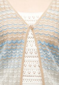 Μπεζ και μπλε ζικ-ζακ πλεκτή ζακέτα, κλειστή με ένα μόνο στρογγυλό κουμπί, πάνω από λευκό πλεκτό τοπ με κροσέ.