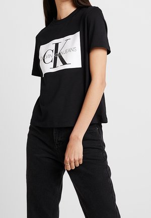 T-shirt en coton noir avec un imprimé rectangulaire blanc portant le texte "CALVIN KLEIN JEANS" et un grand logo "CK". Manches courtes et coupe décontractée.