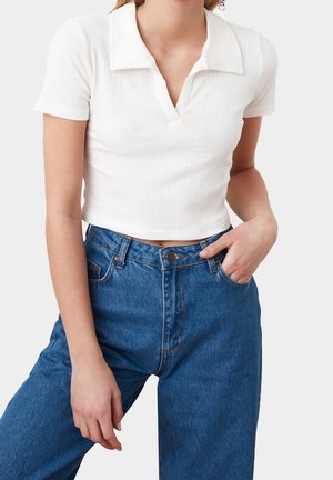 Woman wearing a white short-sleeve ribbed crop top with collar and blue high-waisted jeans, one hand in pocket against plain background.