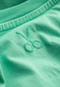 Muntgroene stof met een gestructureerd oppervlak heeft een lichtgroen geborduurd schaarontwerp. De bovenkant heeft een geribde halslijn.