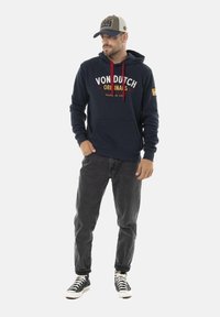 Un sweat à capuche navy avec des cordons de serrage rouges, arborant du texte blanc et jaune. Associé à un jean noir et des baskets noir et blanc. Casquette avec accent bleu.