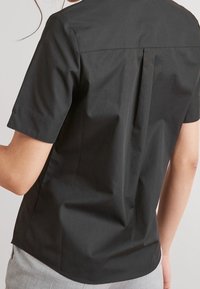 Mustat lyhyet hiharat shirt, jossa on takapleatti, sileä puuvillakangas, pyöristetty helma ja minimalistinen muotoilu. Ei näkyviä logoja tai koristeita.