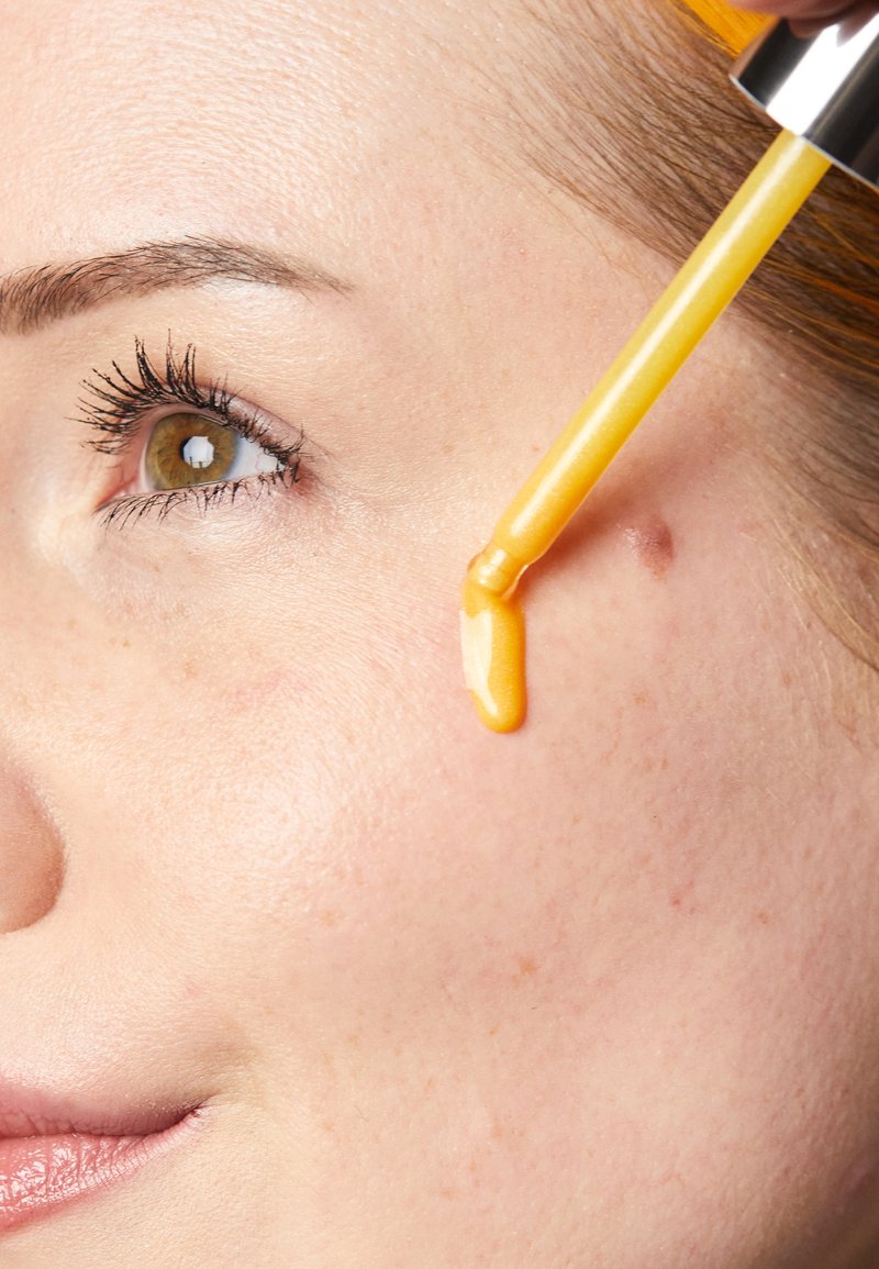 Klar ansiktsserum i en pipett som droppar en gul vätska på ljus, slät hud. Naturliga ögonbryn och ögonfransar ramar in ögat.