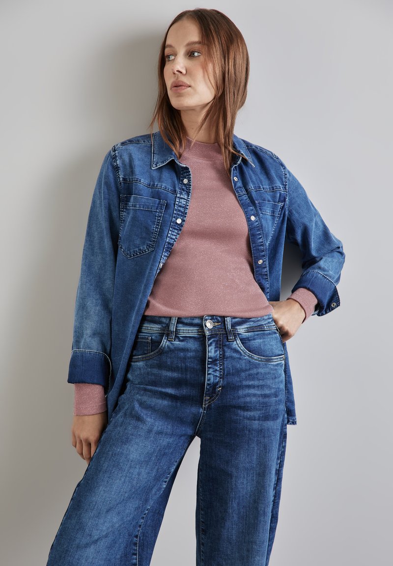 Tummansinisestä denim-paidasta, jossa on kaksi rintataskua, on päällystetty vaaleanpunaisella pitkähihaisella paidalla. Lahkeista leveät siniset farkut, joissa on klassinen malli.