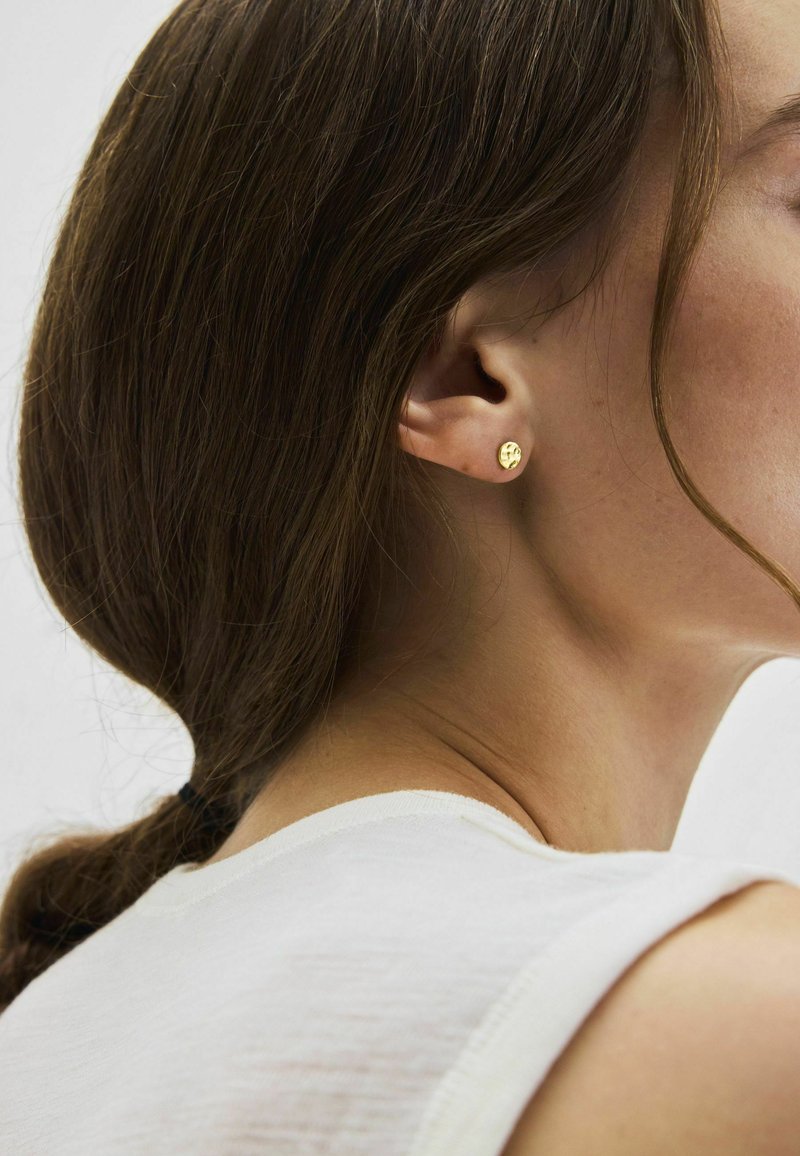Femme aux cheveux bruns attachés en queue de cheval, portant une petite boucle d'oreille ronde en or et un haut blanc sans manches, sur un fond neutre.