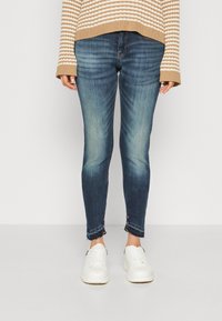 Jeans ajustados en denim azul oscuro con un lavado desgastado, con dobladillos deshilachados y detalles de costura. Combinados con zapatillas blancas y un suéter a rayas.