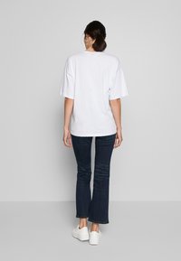 Biała, oversize koszulka z krótkim rękawem, w zestawieniu z ciemnoniebieskimi dzwonami i białymi trampkami, widok z tyłu. Prosty design, miękka tekstura.