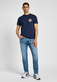 Granatowa bawełniana koszulka z naszywką logo na klatce piersiowej, w połączeniu z jasnoniebieskimi dżinsami i białymi trampkami z czarnymi akcentami.