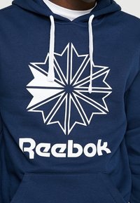 Felpa con cappuccio blu navy con un design geometrico a forma di stella bianca e un grande logo "Reebok" bianco nella parte frontale. Realizzata in un morbido tessuto testurizzato.