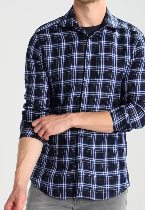 Homme retroussant les manches d'une chemise à carreaux noirs et bleus à boutons, portant un pantalon gris et un sous-vêtement noir, sur un fond uni.