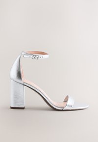 Sandale argentée à talon haut avec bride à la cheville et talon bloc, design à bout ouvert sur fond neutre uni.