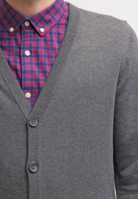 Graue Strickjacke mit V-Ausschnitt und vier Knöpfen, getragen über einem rot-blau karierten Hemd mit Button-Down-Kragen.