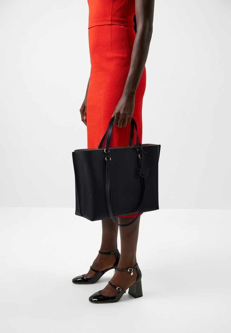 Granatowa skórzana torba na ramię z podwójnymi uchwytami, złotymi akcentami i teksturowanym wykończeniem, trzymana przez osobę ubraną w czerwoną sukienkę i czarne buty.