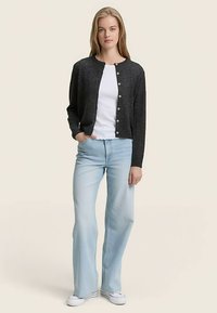 Anglies spalvos kardiganas iš minkštos medžiagos su sagomis priekyje, dėvimas ant baltos palaidinės ir derinamas su šviesiai mėlynos spalvos išplatėjusiais džinsais.