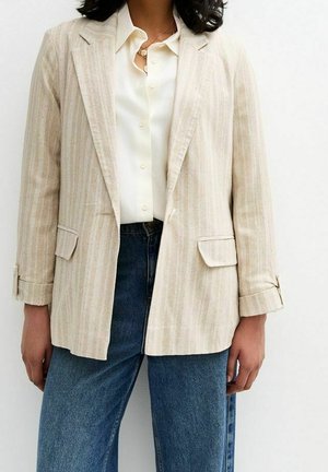 Woman wearing a beige striped blazer over a white button-up shirt paired with blue jeans, standing against a plain light background.