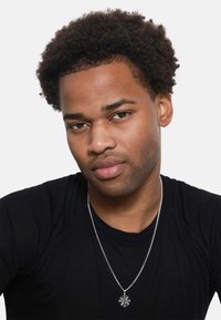 Chemise noire à manches longues ; collier en chaîne argentée avec un pendentif en forme de boussole. Cheveux courts et bouclés et arrière-plan neutre. Gros plan sur les traits du visage.