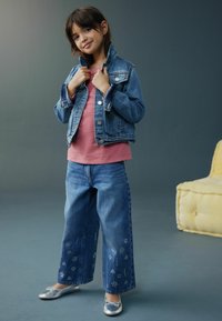 Jachetă din denim peste un top roz, blugi din denim cu croială largă și broderie florală, și pantofi platați. Schema de culori albastru deschis și roz.