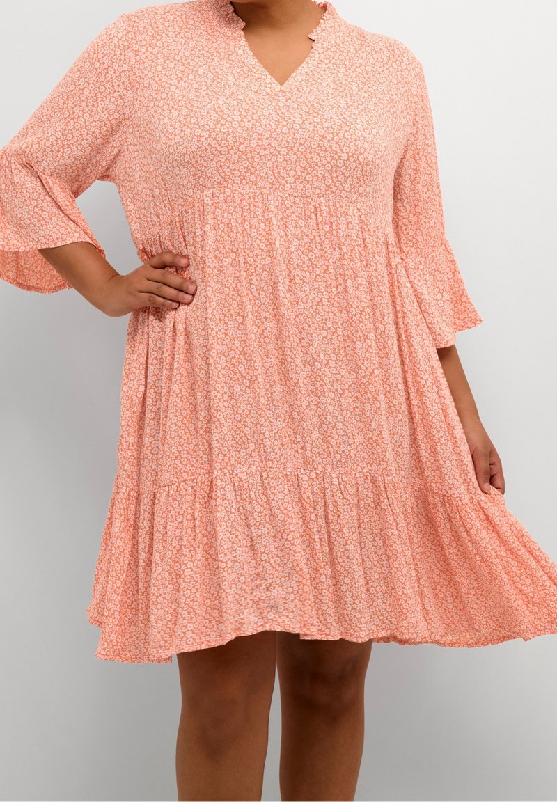 Femme portant une robe ample couleur pêche, longueur genoux, imprimé floral blanc petit, avec des manches cloche trois-quarts, debout devant un fond uni.