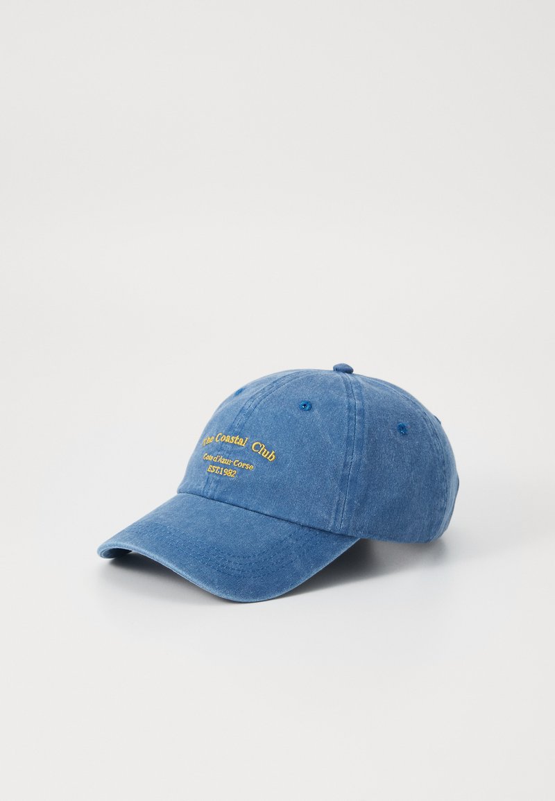 Denim modra kapa za bejzbol z nastavljivim trakom, ki ima rumen vezen napis "The Coastal Club" in ukrivljen rob.