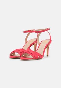 Červené semišové sandály na vysokém podpatku s pásky kolem kotníku a otevřenými prsty, vystavené vedle sebe na bílém pozadí.