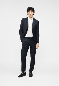 Νέος άνδρας όρθιος, φοράει σκούρο μπλε κοστούμι με λευκό πουκάμισο και μαύρα επίσημα παπούτσια, μπροστά σε απλό φωτεινό φόντο.