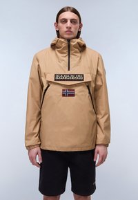 Beige vandtæt jakke med høj krave, forlommer, sorte lynlåse og et norsk flag-patch; parret med sorte shorts.