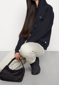 Sweat à capuche noir avec une poche avant, pantalon beige et sac noir. Des baskets noires texturées complètent la tenue, avec des lignes élégantes.