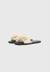 Sandali slide con tomaia in tessuto crema con volant sfrangiati, design open toe e suola in gomma nera. Forma semplice e minimalista.
